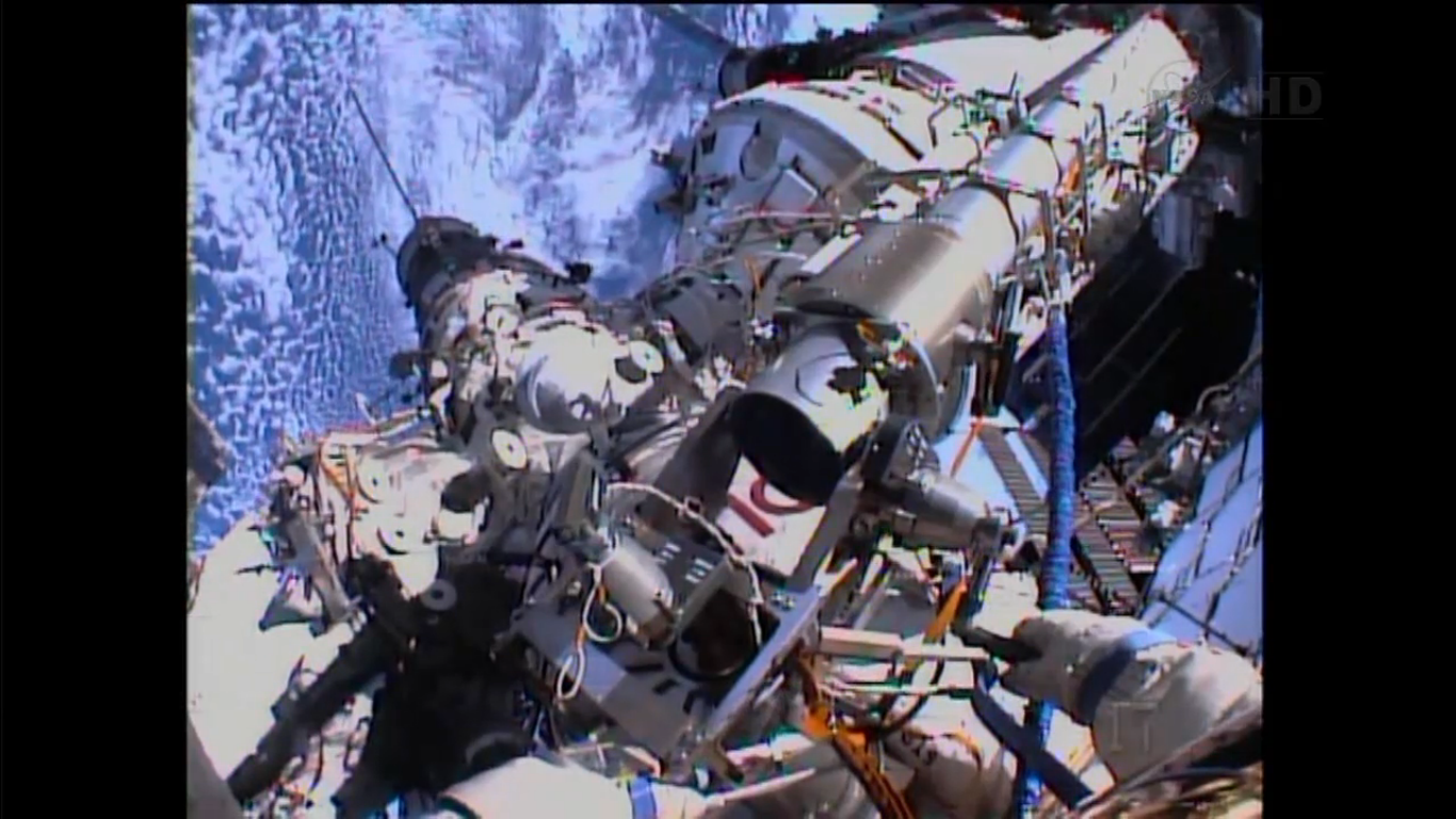 Sasha Mirsukin manouevrant l'un des 2 bras Strela du module russe Pirs lors de l'EVA 34, sous fond de Terre éclairée (capture d'écran)